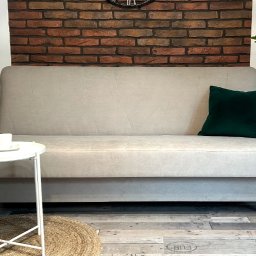 Sebastian Szewczuk - Beżowa sofa z zieloną poduszką na tle ceglanej ściany, uzupełniona roślinami w doniczkach i stolikiem kawowym z filiżanką, tworząca przytulną aranżację wnętrza.