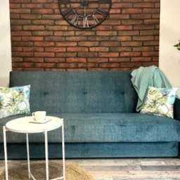 Turkusowa sofa z poduszkami w tropikalne wzory na tle ściany z cegły, uzupełniona stolikiem i roślinami doniczkowymi, tworzy przytulny kącik wypoczynkowy.