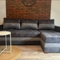 Szara, tapicerowana sofa narożna z pufą i okrągłym stolikiem na tle ceglanej ściany z zegarem, uzupełniona roślinami doniczkowymi i jutowym dywanem.