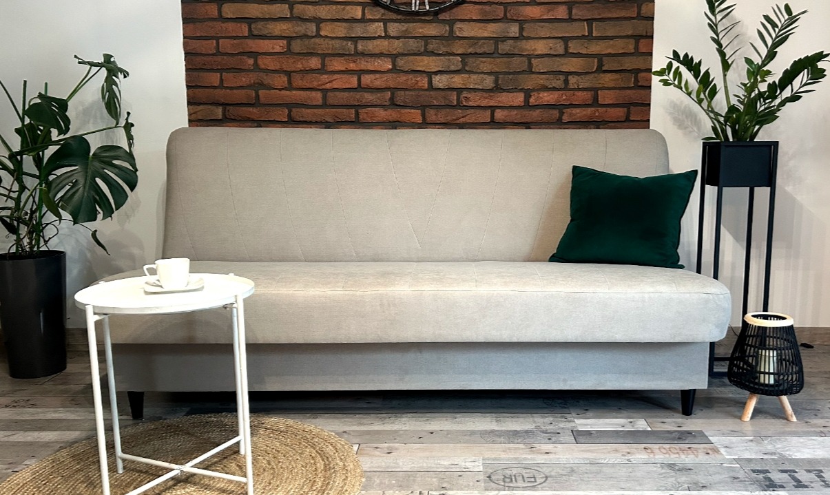 Beżowa sofa z zieloną poduszką na tle ceglanej ściany, uzupełniona roślinami w doniczkach i stolikiem kawowym z filiżanką, tworząca przytulną aranżację wnętrza.