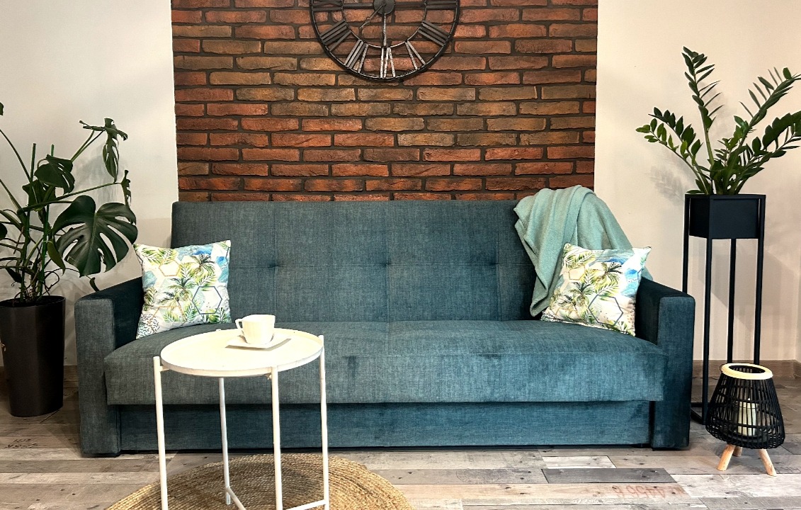 Turkusowa sofa z poduszkami w tropikalne wzory na tle ściany z cegły, uzupełniona stolikiem i roślinami doniczkowymi, tworzy przytulny kącik wypoczynkowy.