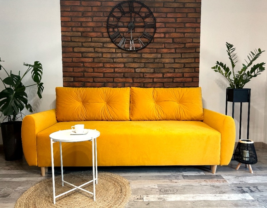 Żółta sofa z poduszkami na drewnianych nóżkach, stolik z filiżanką, zegar na ceglanej ścianie, rośliny w donicach, dywanik na drewnianej podłodze.