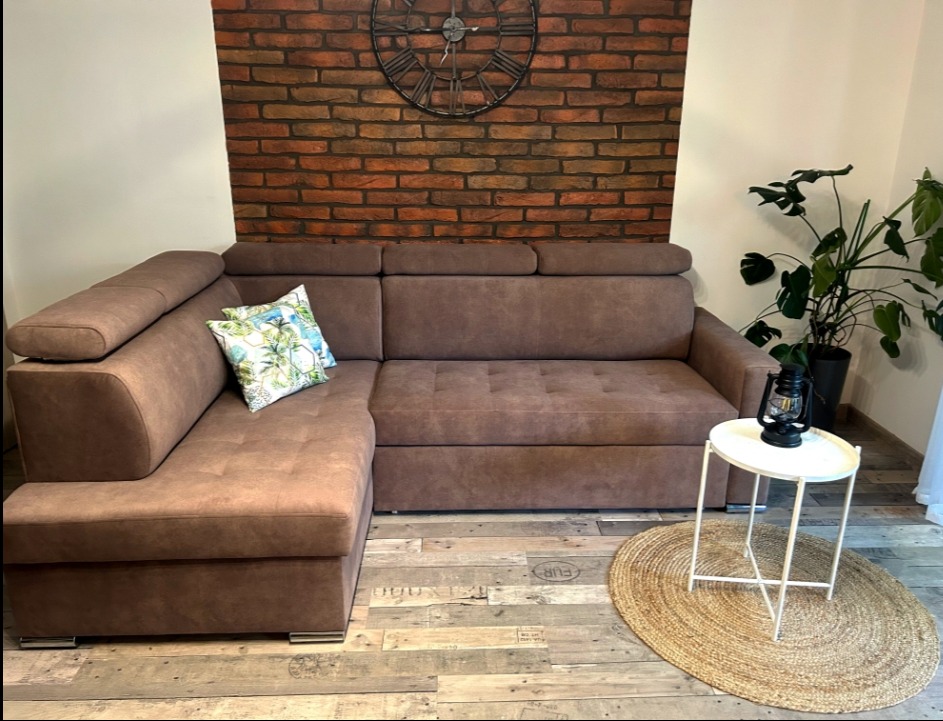 Brązowy narożnik z funkcją spania, poduszkami i zegarem na ceglanej ścianie. Obok stolik z lampą na tle rośliny doniczkowej.