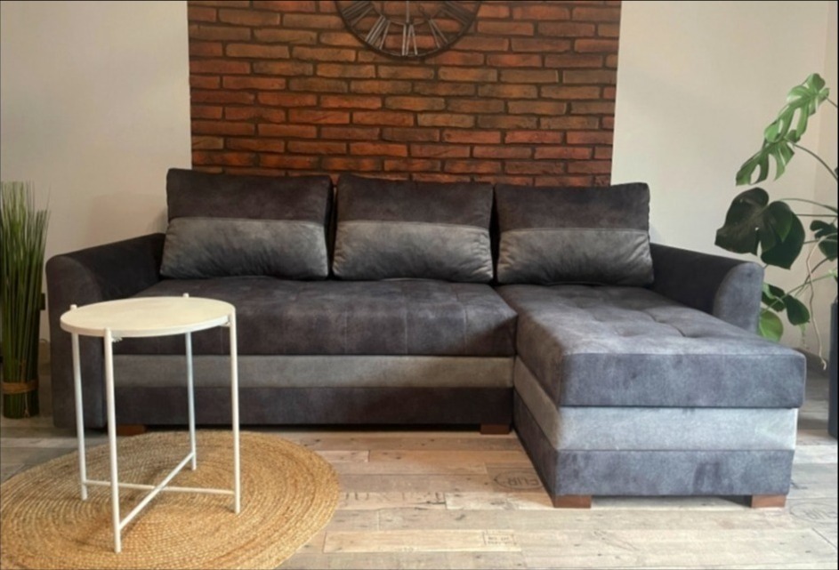 Szara, tapicerowana sofa narożna z pufą i okrągłym stolikiem na tle ceglanej ściany z zegarem, uzupełniona roślinami doniczkowymi i jutowym dywanem.