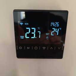 Piotr Mogilski - Nowoczesny, czarny termostat pokojowy z wyświetlaną temperaturą 23.1°C i ustawioną na 24°C, zamontowany na jasnej ścianie. Widoczne ikony ustawień i godzina 14:26.