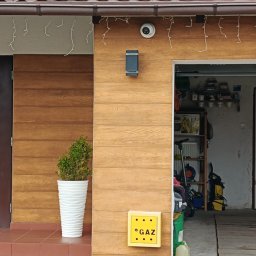 Piotr Mogilski - Elewacja domu z drewnianą okładziną, oświetleniem zewnętrznym, kamerą i skrzynką gazową. Widoczne dekoracyjne lampki i wejście do garażu.