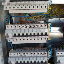 Piotr Mogilski - Rozdzielnica elektryczna z zainstalowanymi bezpiecznikami Legrand, widoczne przewody i oznaczenia obwodów. Instalacja w szarej metalowej obudowie.