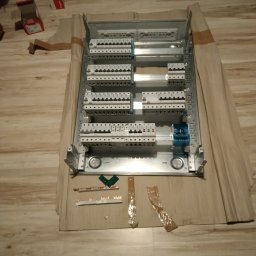 Piotr Mogilski - Rozdzielnica elektryczna w trakcie montażu, widoczne zabezpieczenia, przewody i elementy montażowe. Rozłożona na kartonie na podłodze imitującej drewno.