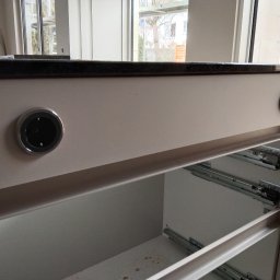 Piotr Mogilski - Szary blat z gniazdem elektrycznym i otworem na kabel, widoczne szuflady z metalowymi prowadnicami. Nowoczesny design, jasne wnętrze, minimalistyczny styl.