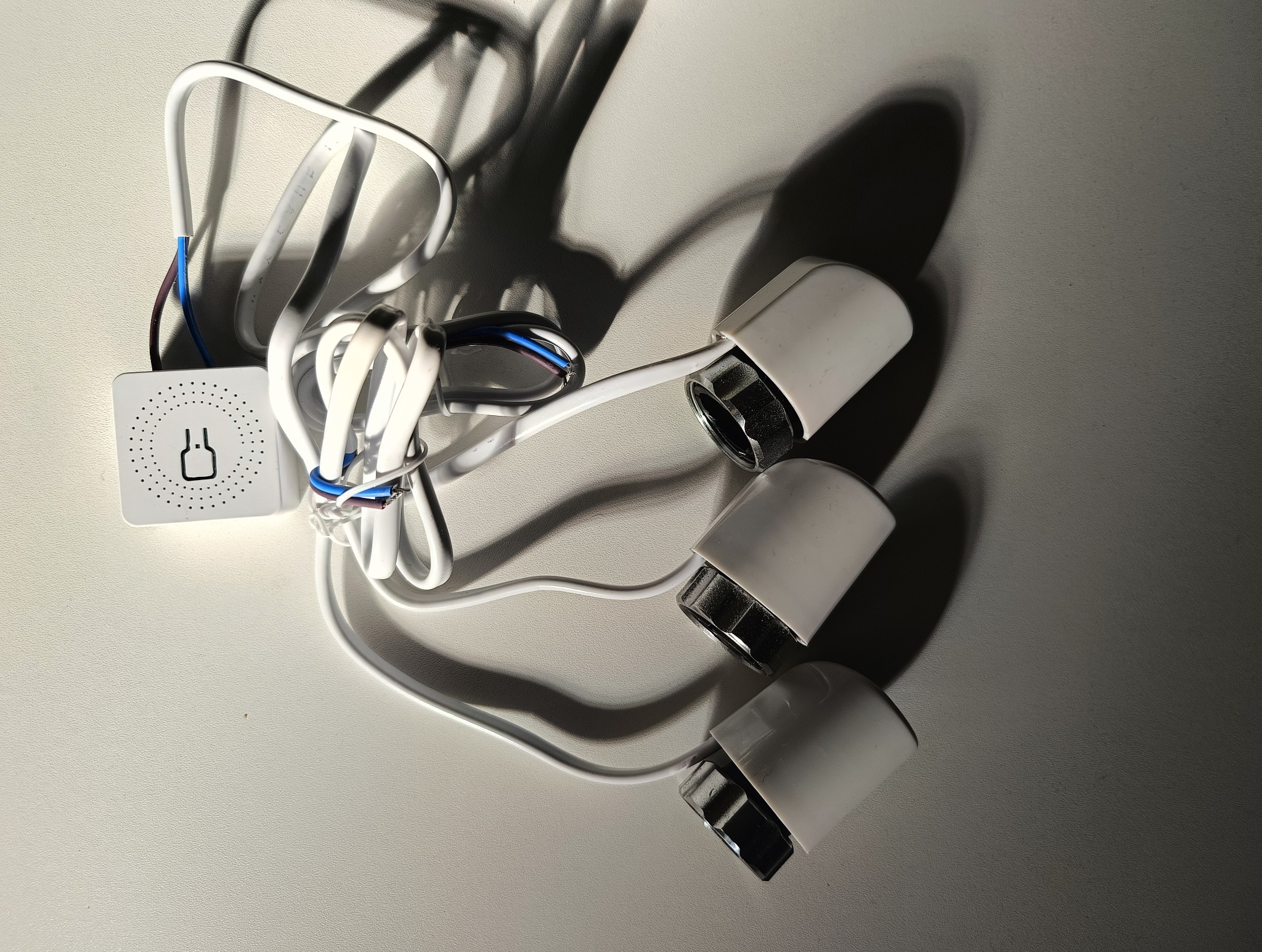 Biały moduł sterujący z trzema podłączonymi głowicami termostatycznymi i przewodami na jasnym tle, oświetlony z góry.