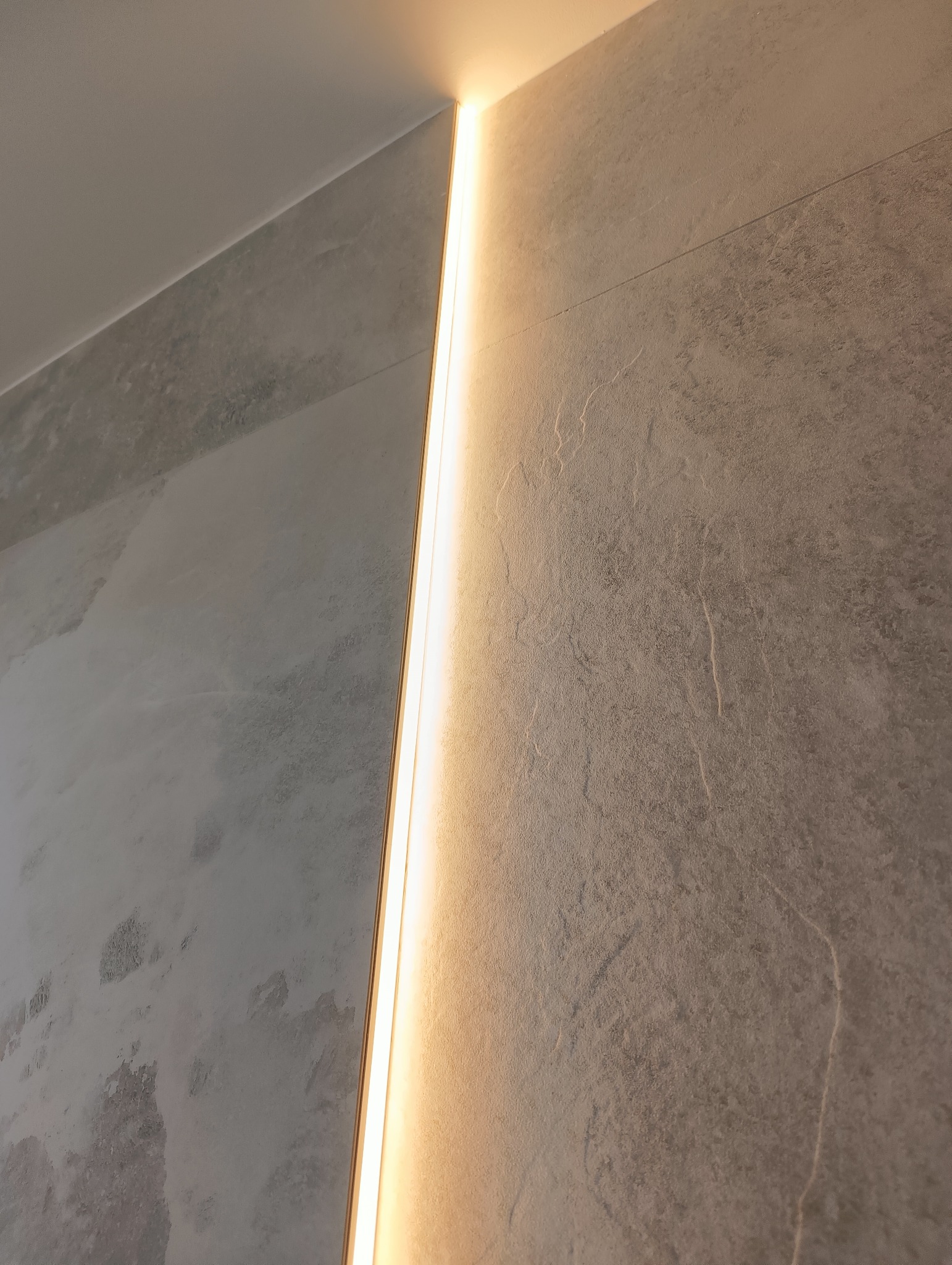 Wąska listwa LED, pionowo wkomponowana w narożnik ściany wykończonej szarą, strukturalną płytą. Ciepłe światło podkreśla fakturę materiału.