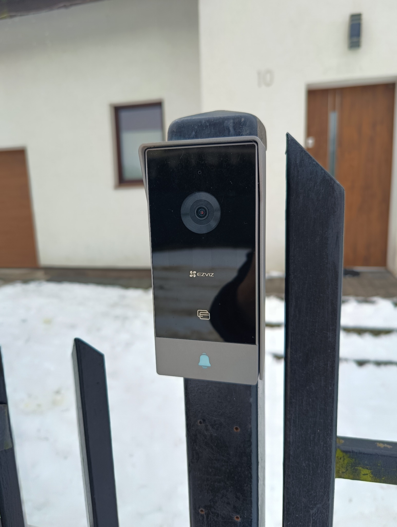Nowoczesny wideodomofon marki EZVIZ zamontowany na czarnym, metalowym słupku ogrodzenia posesji. W tle biały budynek z numerem 10 i brązowymi drzwiami wejściowymi.