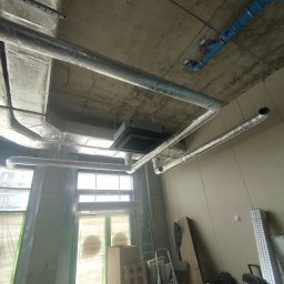INSTAL-KOM Mariusz Suknarowski - Instalacja wentylacyjna z srebrnymi rurami i niebieskimi elementami, widoczna pod betonowym sufitem w surowym wnętrzu budynku w trakcie budowy.
