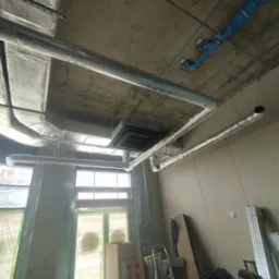 Instalacja wentylacyjna z srebrnymi rurami i niebieskimi elementami, widoczna pod betonowym sufitem w surowym wnętrzu budynku w trakcie budowy.