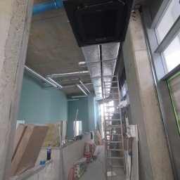 INSTAL-KOM Mariusz Suknarowski - Montaż klimatyzacji kasetonowej w surowym wnętrzu. Widoczne rury wentylacyjne, drabina i materiały budowlane. Ujęcie z dołu, podkreślające wysokość pomieszczenia.