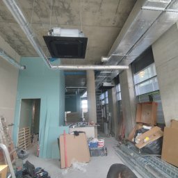 INSTAL-KOM Mariusz Suknarowski - Montaż klimatyzacji sufitowej w surowym wnętrzu. Widoczne kanały wentylacyjne, narzędzia i osoba pracująca przy instalacji. Stan surowy, płyty gipsowe.