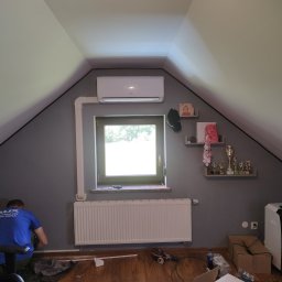 INSTAL-KOM Mariusz Suknarowski - Montaż klimatyzacji w pokoju na poddaszu: biały klimatyzator nad oknem, instalacja w listwach, grzejnik pod oknem, instalator w niebieskiej koszulce przy ścianie.