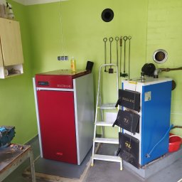 INSTAL-KOM Mariusz Suknarowski - Dwa kotły grzewcze: czerwony, nowoczesny MCZ Selecta 15 HQ i niebieski, tradycyjny Zębiec, stojące obok drabiny w kotłowni z zielonymi ścianami i narzędziami kominiarskimi.