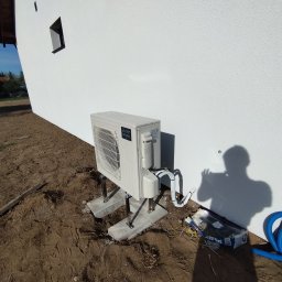 INSTAL-KOM Mariusz Suknarowski - Biała jednostka zewnętrzna pompy ciepła Mitsubishi Ecodan, zamontowana na betonowych podstawach przy ścianie budynku z widocznymi przewodami i elementami instalacji.