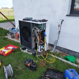 INSTAL-KOM Mariusz Suknarowski - Pompa ciepła w trakcie serwisu na trawie, widoczne narzędzia i otwarte obudowy. Ściana budynku z oknem w tle. Przewody i manometry podłączone.