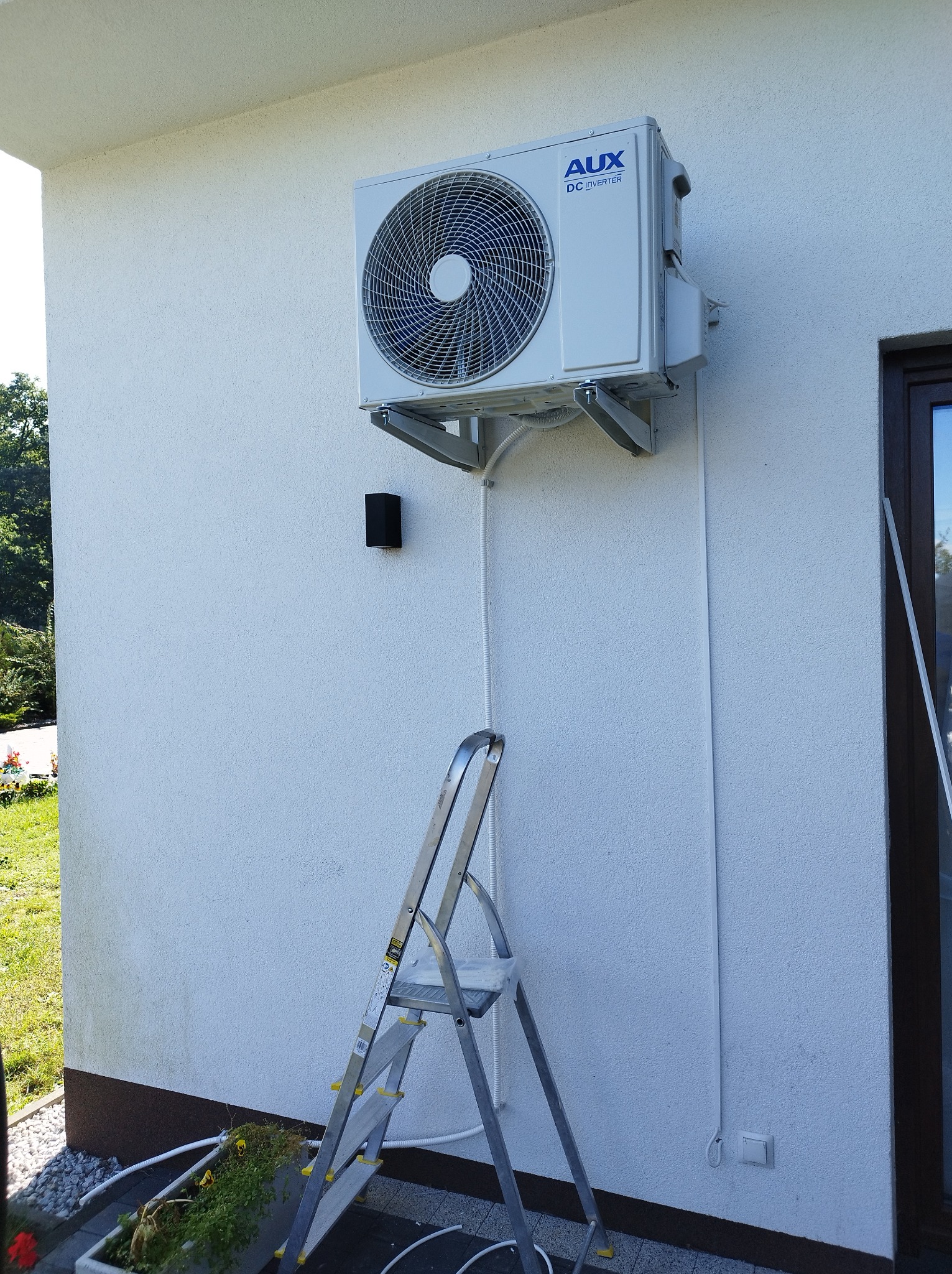 Biała jednostka zewnętrzna klimatyzacji AUX DC Inverter zamontowana na wspornikach na elewacji budynku, obok drabina. Widoczne przewody i lampa elewacyjna.