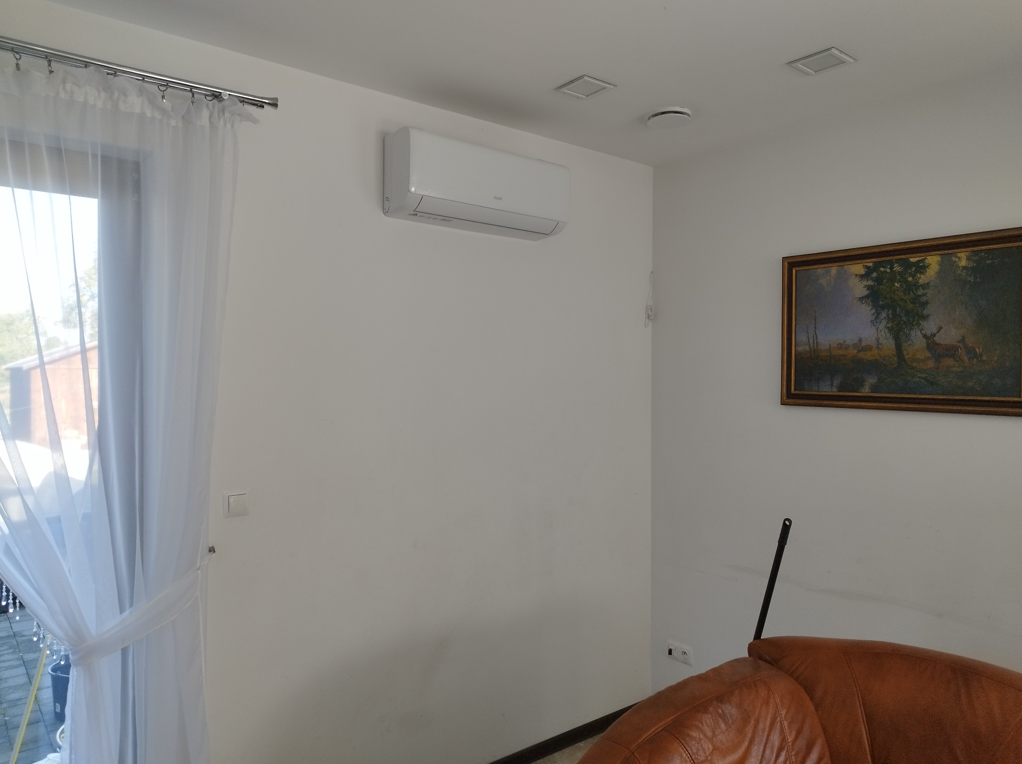 Biały klimatyzator ścienny AUX w salonie, Żary. Widoczne zasłony, obraz z motywem jelenia i fragment skórzanego fotela. Nowoczesny design.