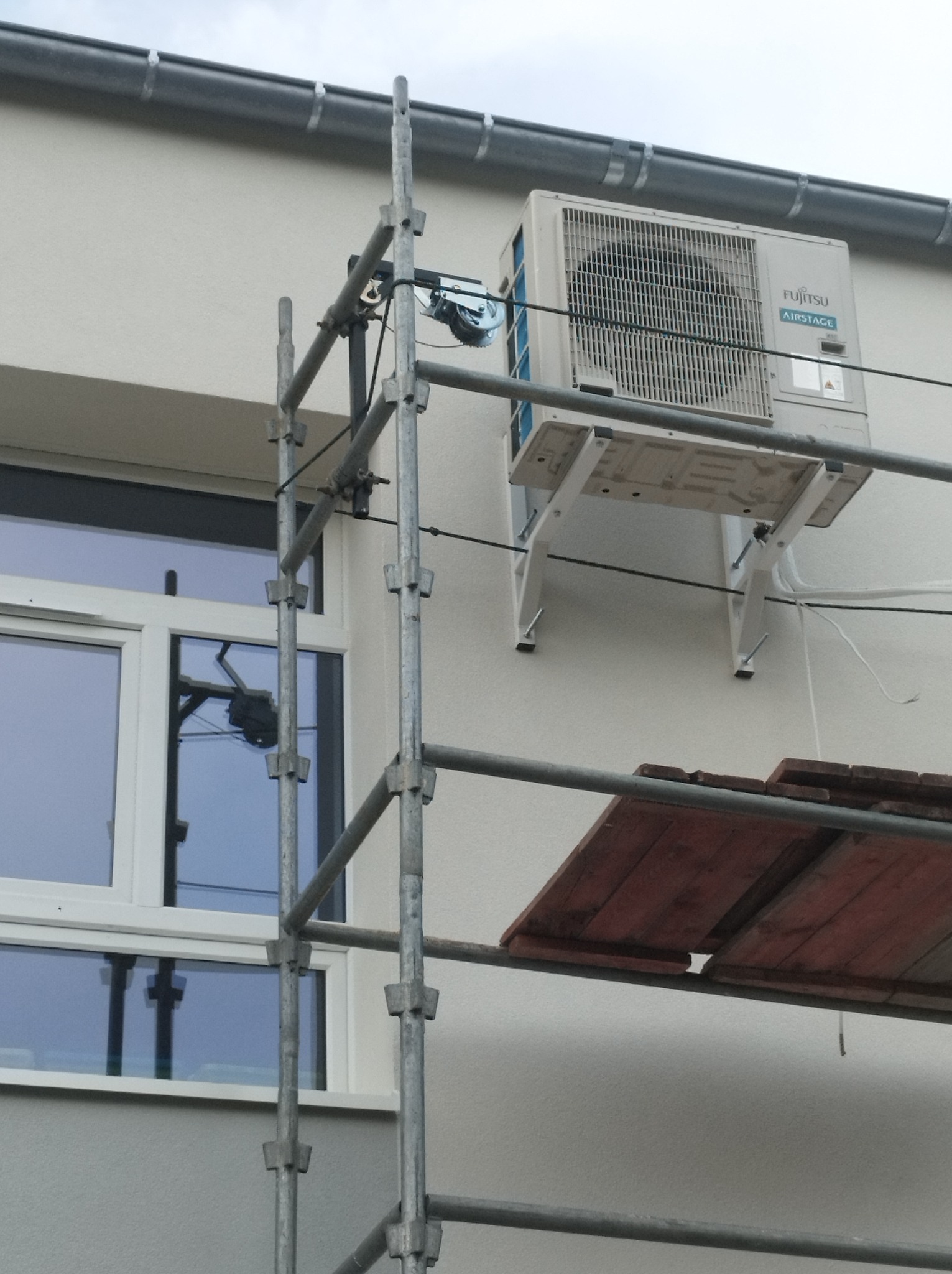 Biała jednostka zewnętrzna klimatyzacji Fujitsu zamontowana na ścianie budynku, widoczna z rusztowania. Okno w tle.
