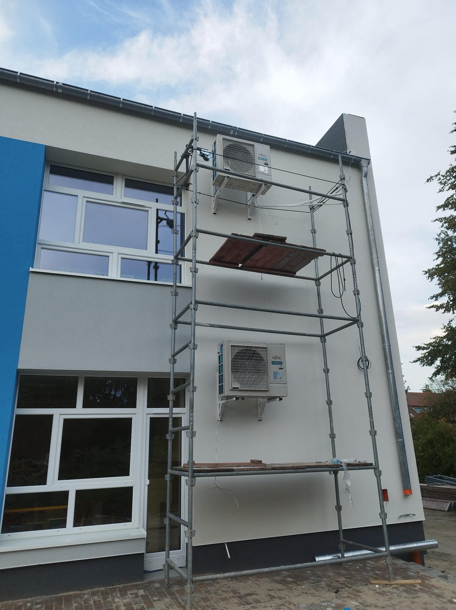 Montaż dwóch klimatyzatorów Fujitsu Airstage na elewacji budynku z pomocą rusztowania. Widoczne okna i rynna.