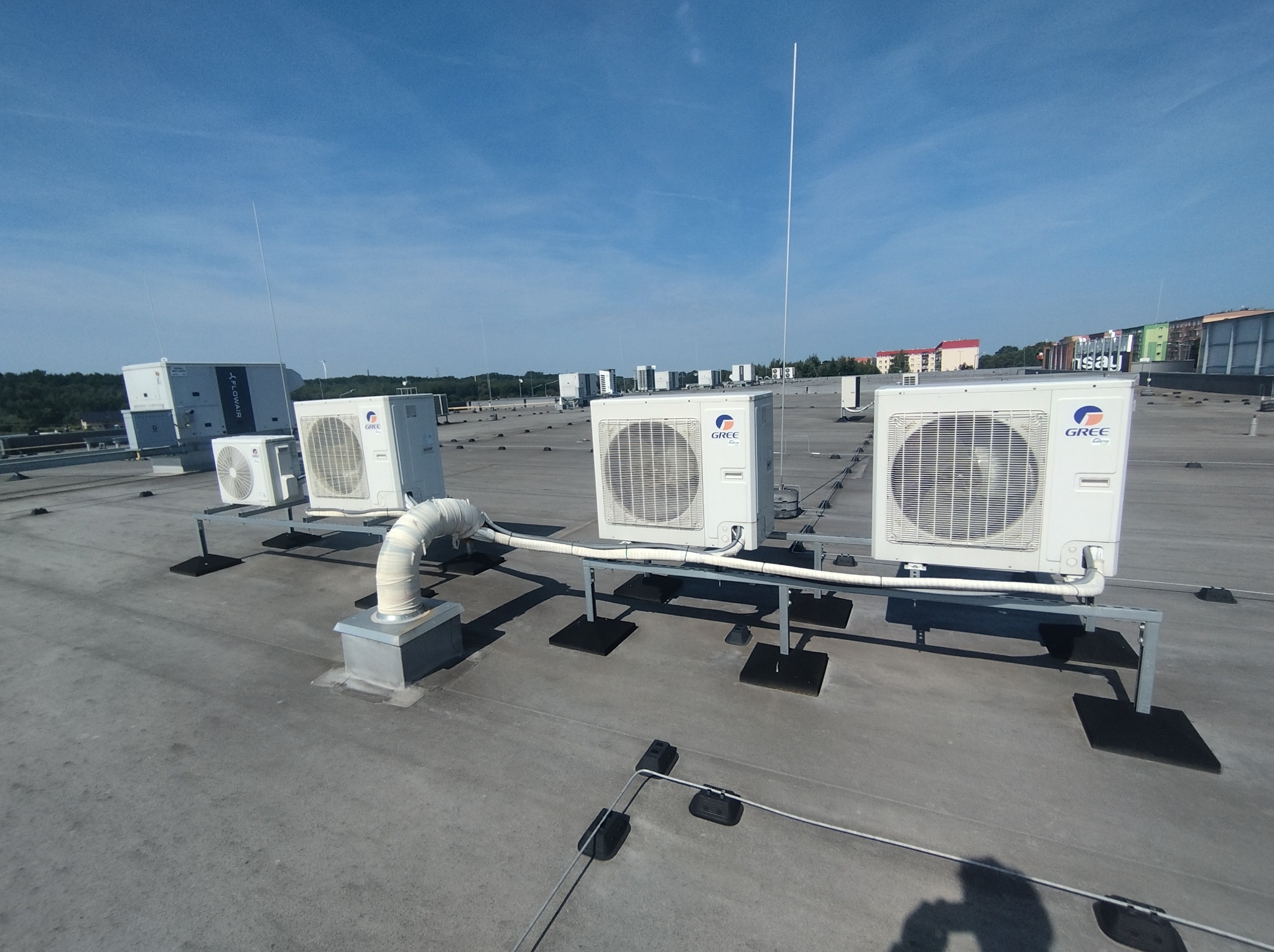 Białe jednostki zewnętrzne klimatyzacji marki Gree, zamontowane na dachu budynku. Widoczne elementy instalacji i tło z budynkami w oddali. Słoneczny dzień.