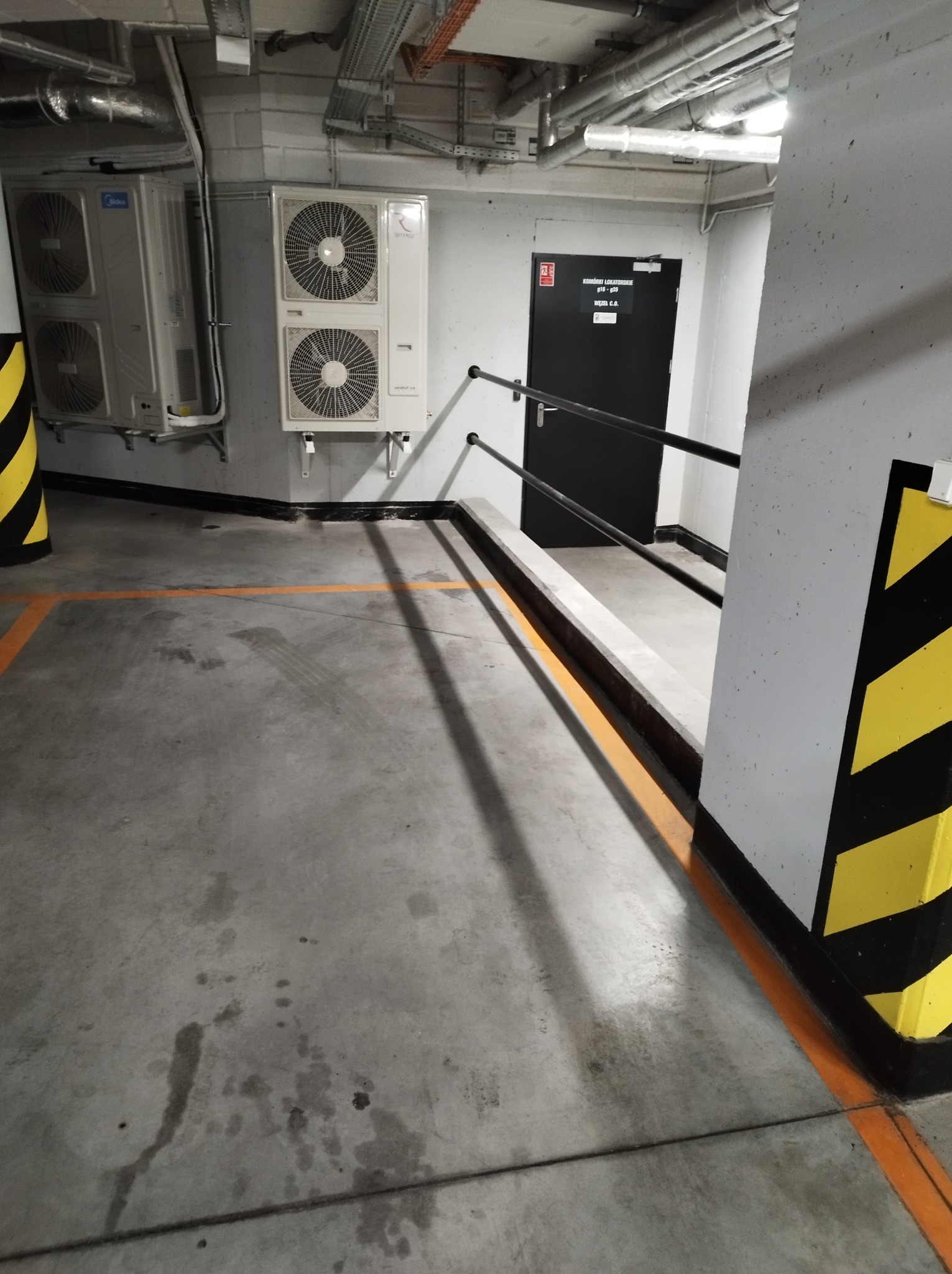 Klimatyzatory Midea i Rotenso zamontowane w garażu podziemnym, widoczne rury instalacyjne pod sufitem i oznaczenia stref żółto-czarnymi pasami.