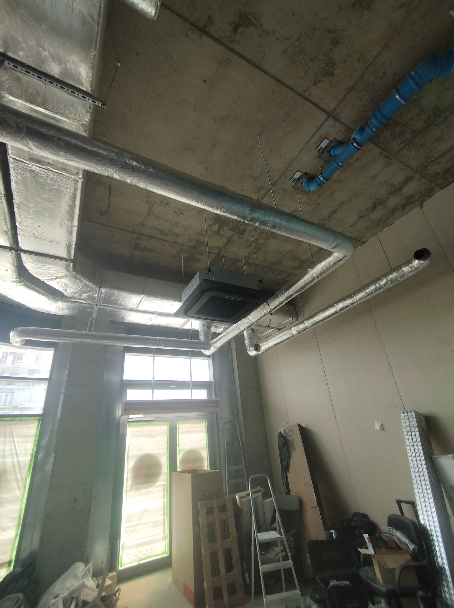 Instalacja wentylacyjna z srebrnymi rurami i niebieskimi elementami, widoczna pod betonowym sufitem w surowym wnętrzu budynku w trakcie budowy.