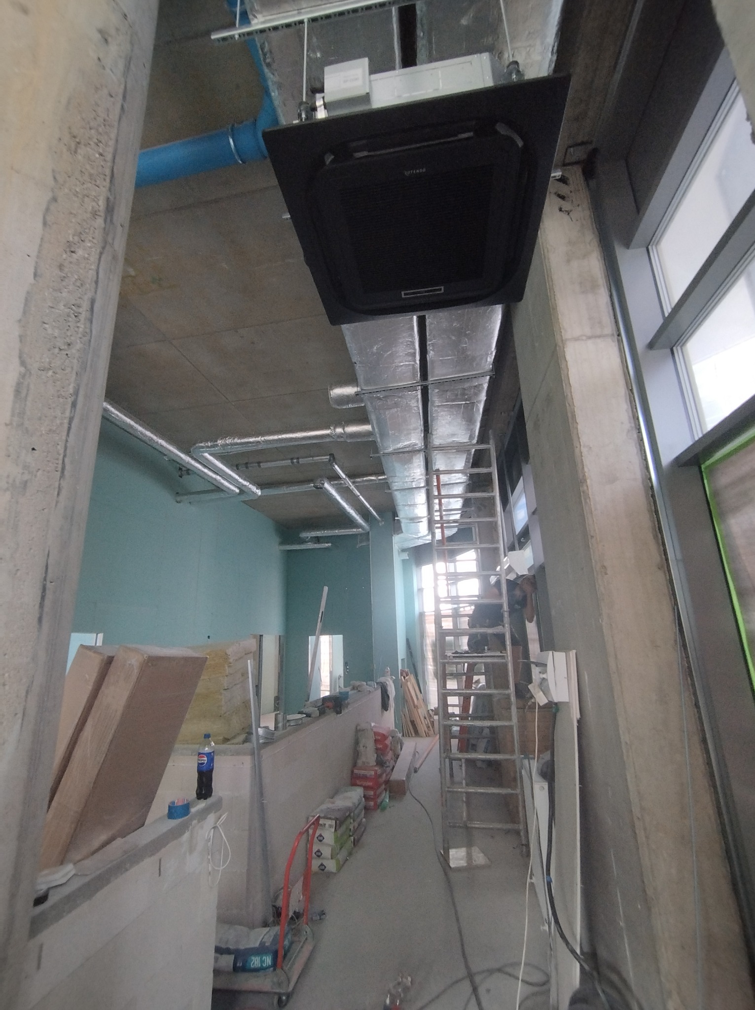 Montaż klimatyzacji kasetonowej w surowym wnętrzu. Widoczne rury wentylacyjne, drabina i materiały budowlane. Ujęcie z dołu, podkreślające wysokość pomieszczenia.