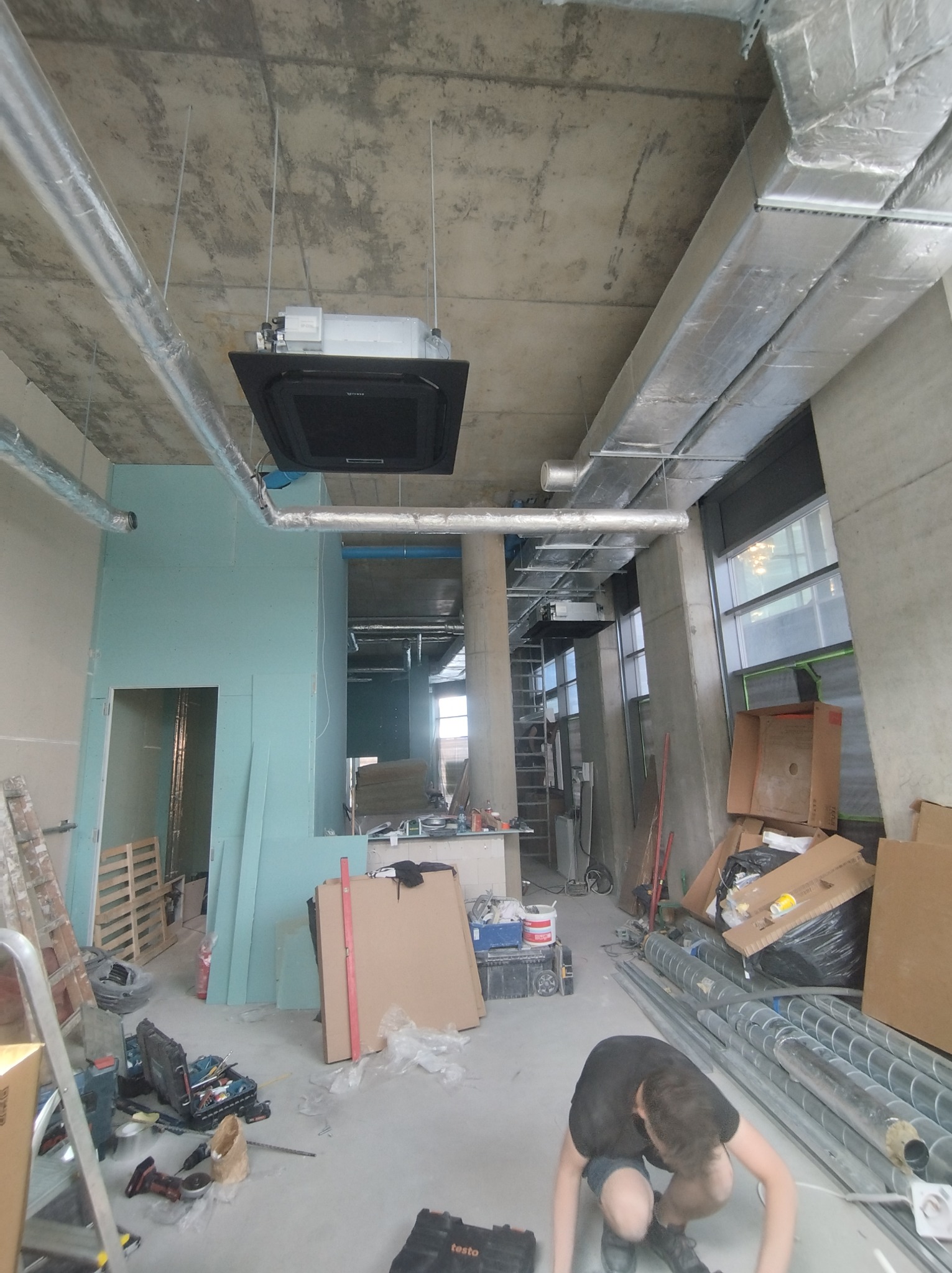 Montaż klimatyzacji sufitowej w surowym wnętrzu. Widoczne kanały wentylacyjne, narzędzia i osoba pracująca przy instalacji. Stan surowy, płyty gipsowe.