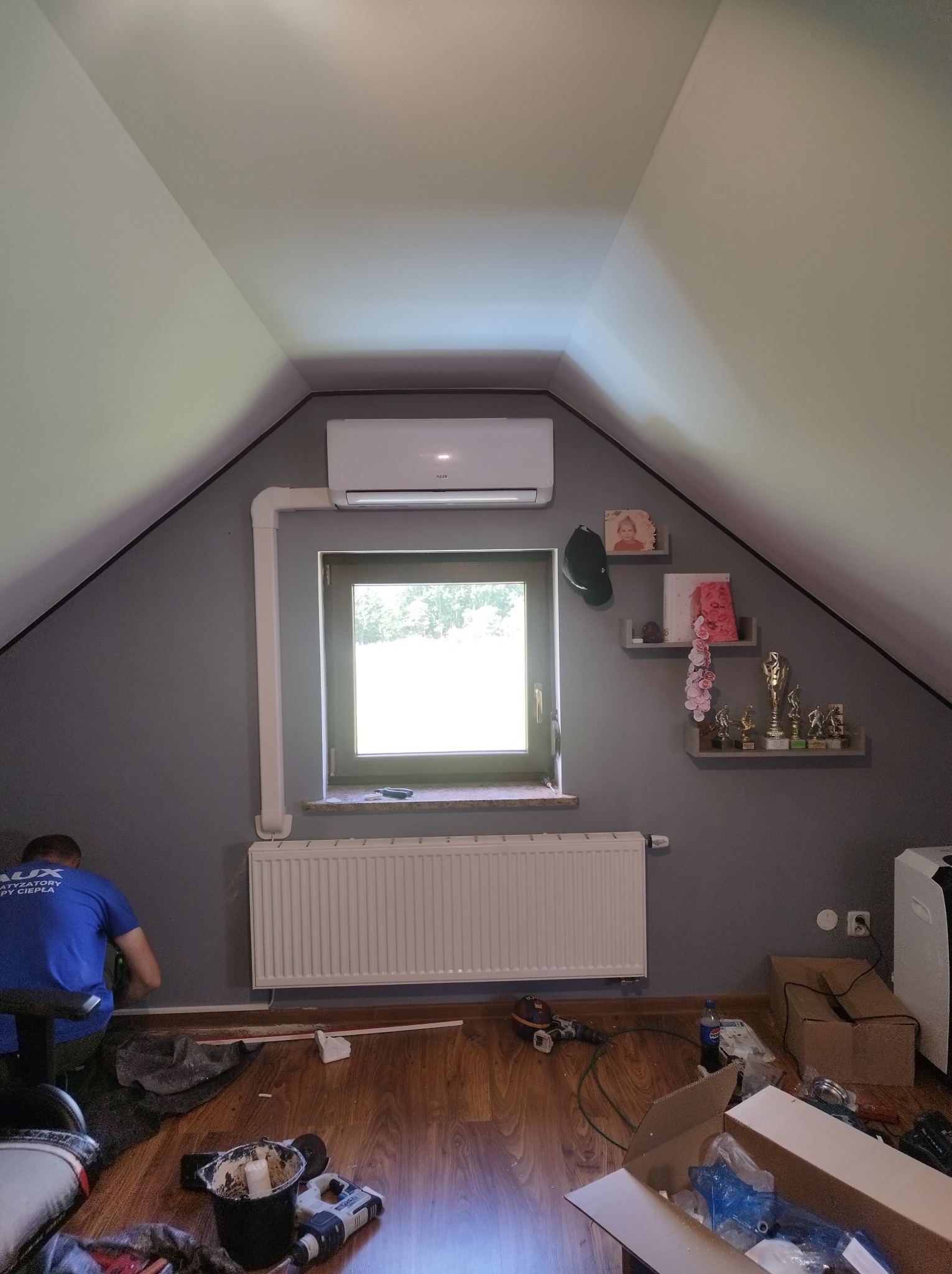 Montaż klimatyzacji w pokoju na poddaszu: biały klimatyzator nad oknem, instalacja w listwach, grzejnik pod oknem, instalator w niebieskiej koszulce przy ścianie.
