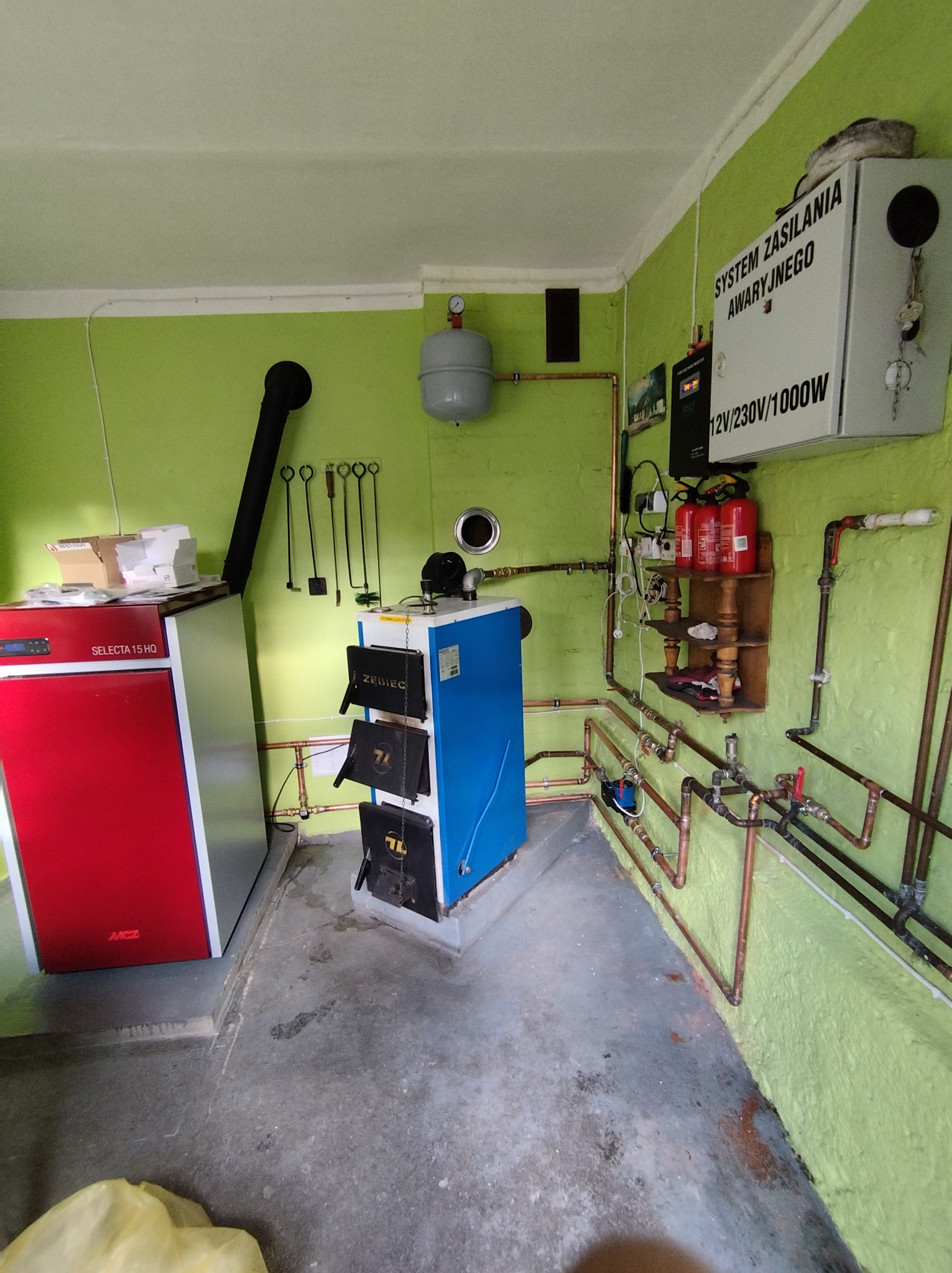 Kotłownia z piecem na paliwo stałe Zębiec i piecem Selecta 15 HQ. Widoczne rury instalacji, zbiornik wyrównawczy i system zasilania awaryjnego. Zielone ściany.
