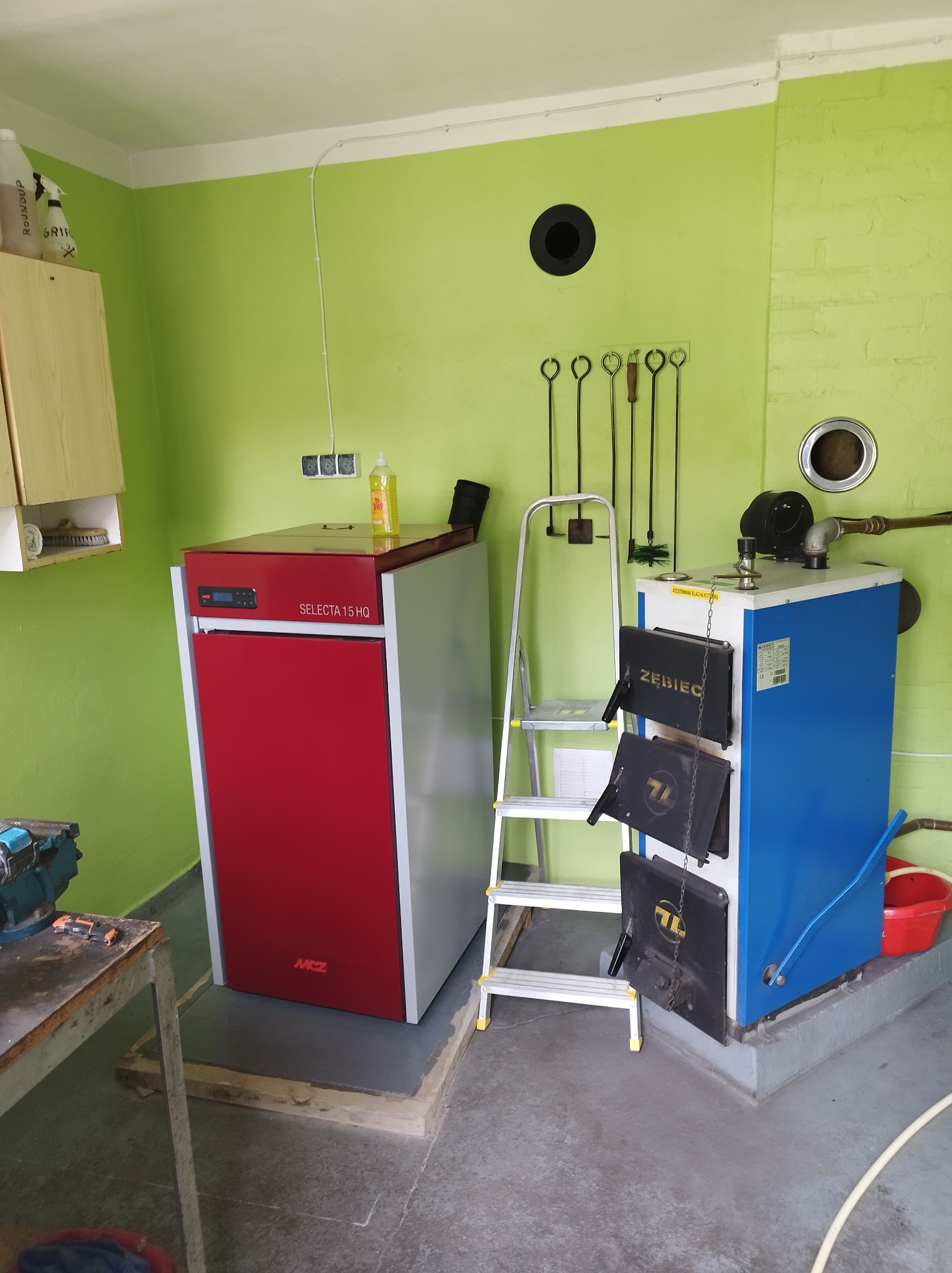 Dwa kotły grzewcze: czerwony, nowoczesny MCZ Selecta 15 HQ i niebieski, tradycyjny Zębiec, stojące obok drabiny w kotłowni z zielonymi ścianami i narzędziami kominiarskimi.