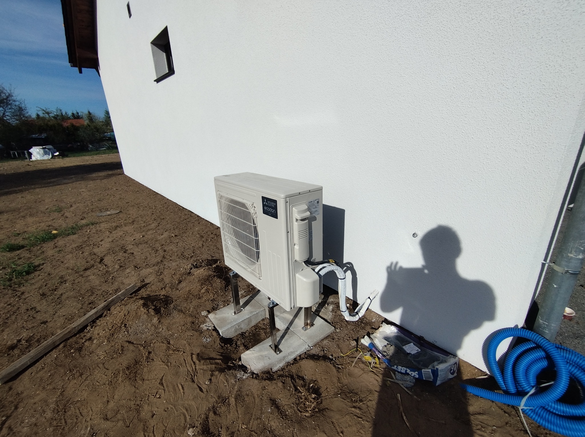 Biała jednostka zewnętrzna pompy ciepła Mitsubishi Ecodan, zamontowana na betonowych podstawach przy ścianie budynku z widocznymi przewodami i elementami instalacji.