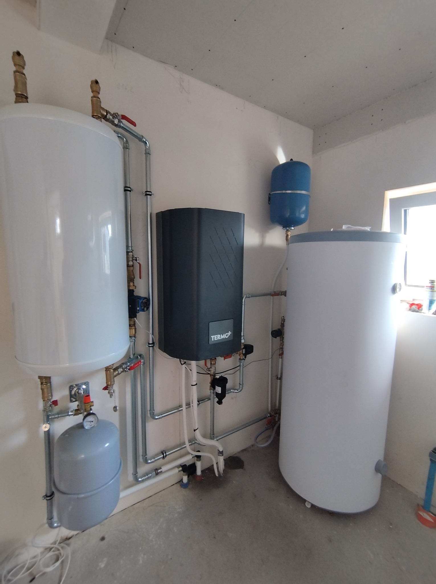 Zestaw urządzeń grzewczych: dwa białe zasobniki ciepłej wody, szary piec TERMO+, niebieski zbiornik wyrównawczy, rury i zawory w surowym pomieszczeniu.