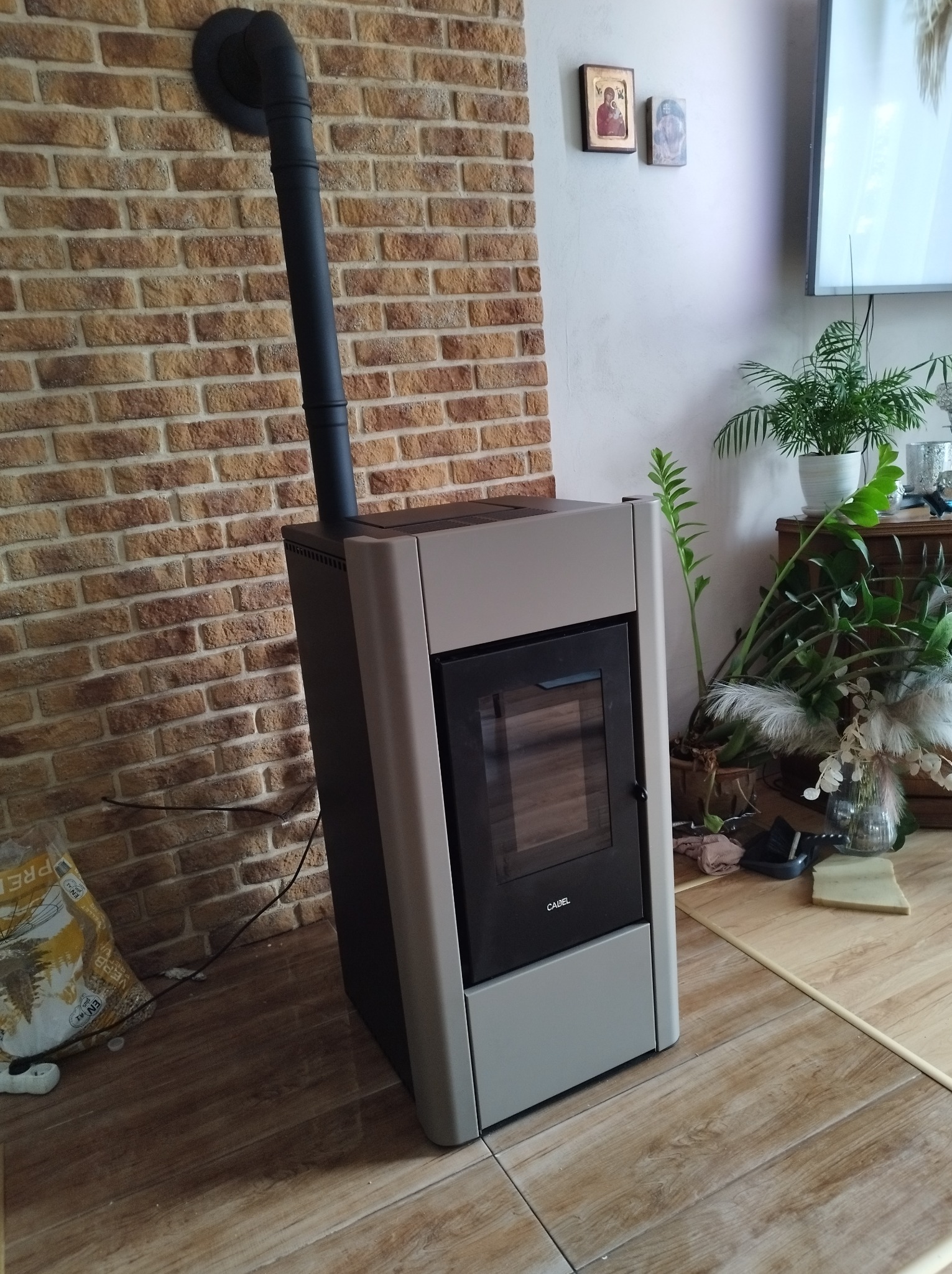 Nowoczesny piec wolnostojący marki CADEL z czarnym kominem, ustawiony na drewnopodobnej podłodze przy ceglanej ścianie w jasnym, domowym wnętrzu.