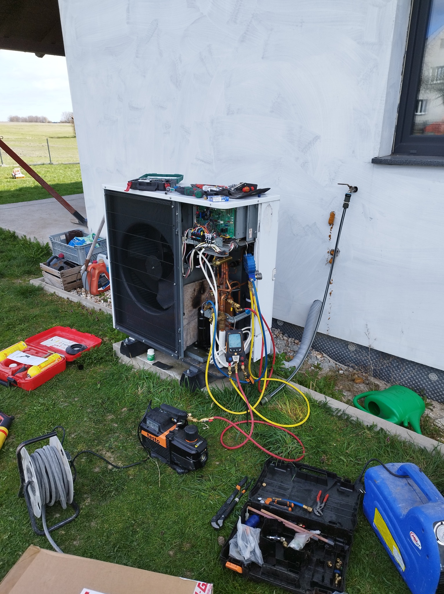Pompa ciepła w trakcie serwisu na trawie, widoczne narzędzia i otwarte obudowy. Ściana budynku z oknem w tle. Przewody i manometry podłączone.