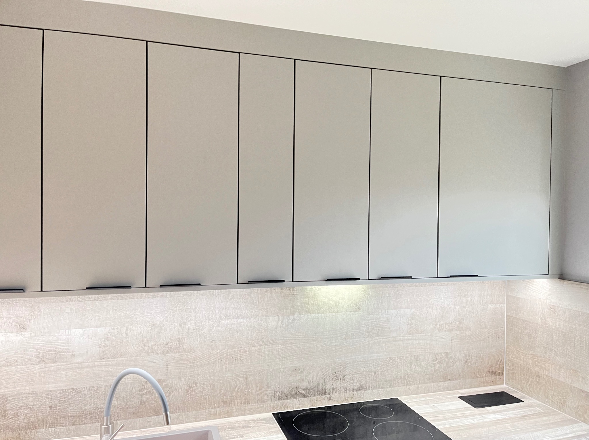 Szare, minimalistyczne szafki kuchenne wiszące nad blatem z płyty imitującej drewno, z widoczną płytą indukcyjną i zlewem. Proste, czarne uchwyty.