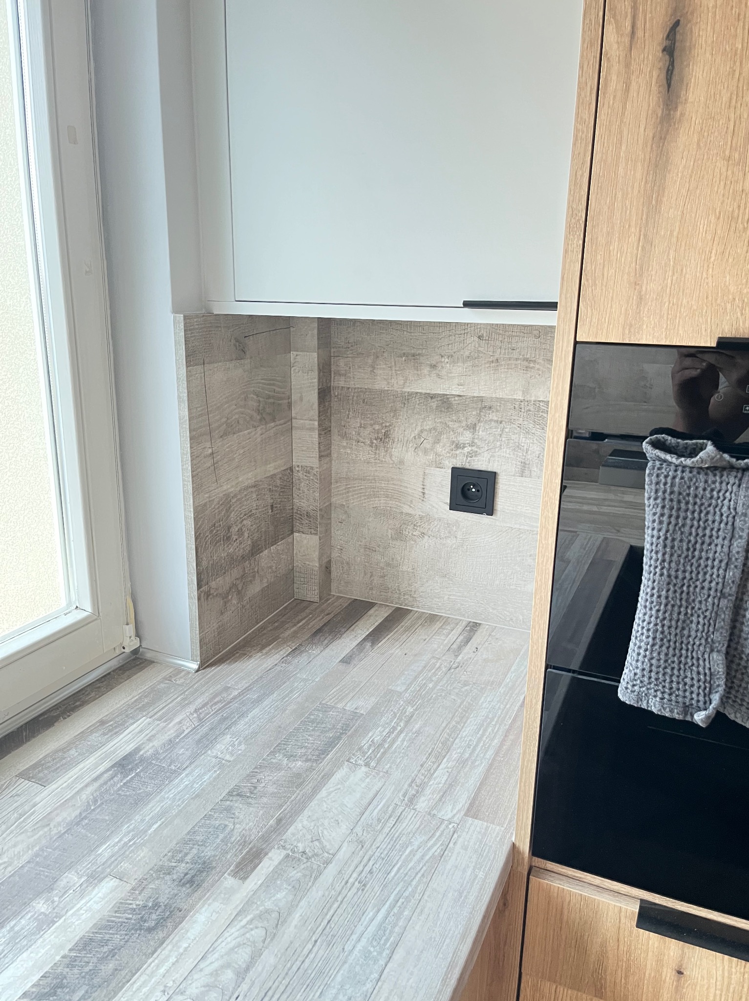 Fragment kuchni z drewnopodobnymi panelami na podłodze i ścianie, czarne gniazdko elektryczne, zabudowa kuchenna z drewnianymi frontami i szarą ścierką.