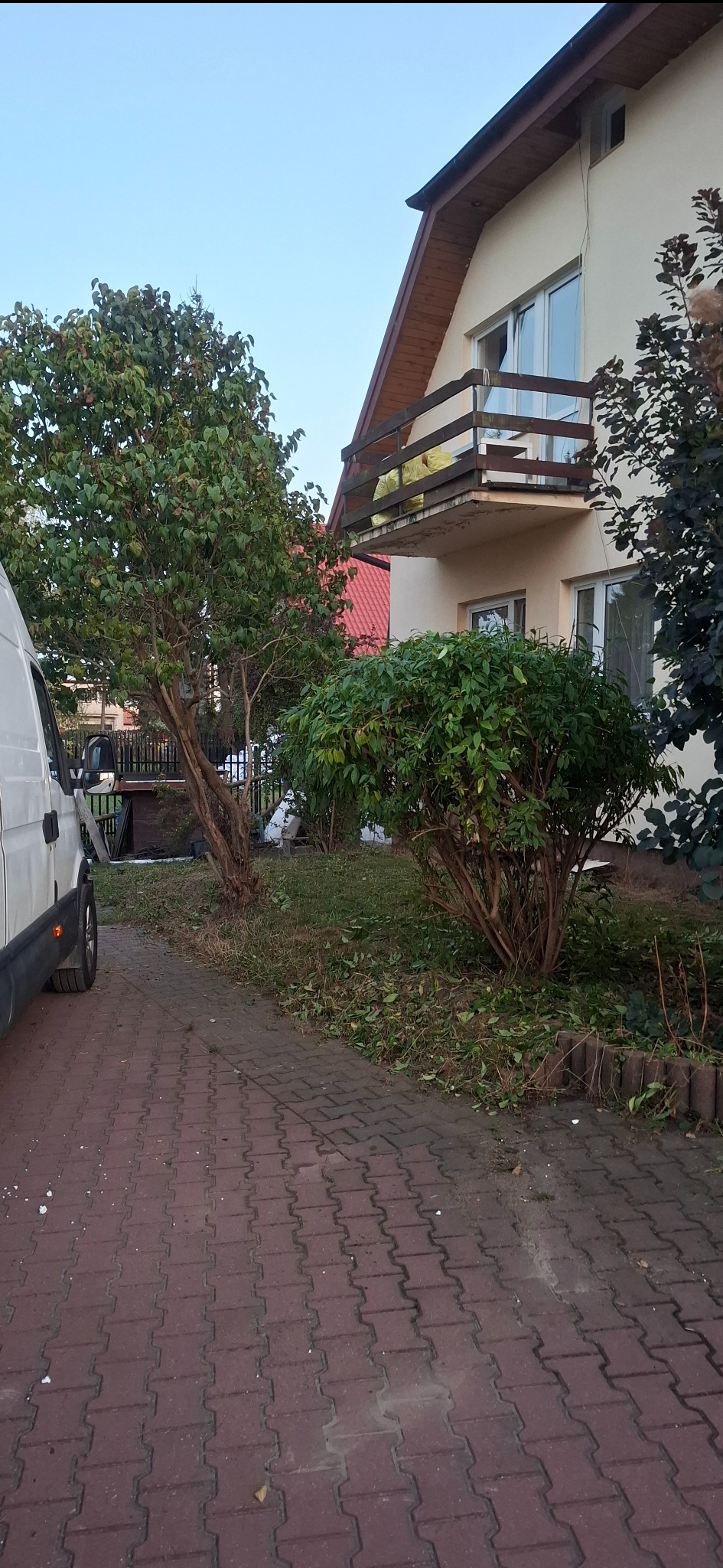 Ogród po przycięciu krzewów i drzew, z widocznymi gałęziami na trawie, obok brukowanej ścieżki i zaparkowanego białego busa. Dom z balkonem w tle.