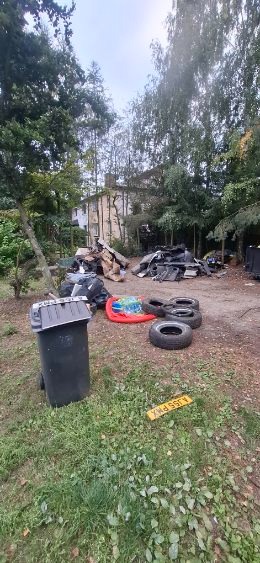 Zabrudzony teren z oponami, elementami karoserii i koszem na śmieci na tle budynku w otoczeniu drzew. Na trawie widoczna żółta tablica rejestracyjna.