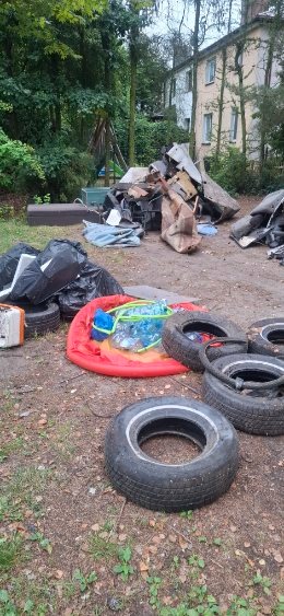 Stos opon, worki ze śmieciami i gruz na zewnątrz, w tle drzewa i budynek. Widoczny nieporządek i pozostałości po pracach porządkowych.