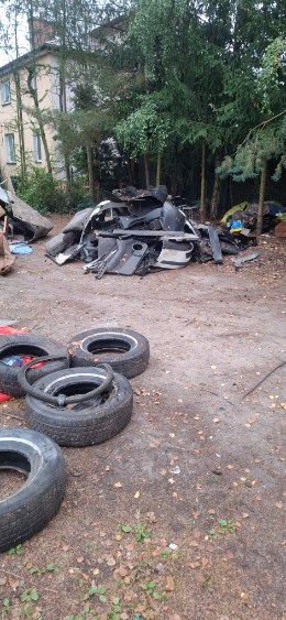 Sterta pociętych części samochodowych i opon na ziemi, w tle budynek i drzewa. Widoczne elementy karoserii, wnętrza pojazdu. Złomowisko, demontaż pojazdów.