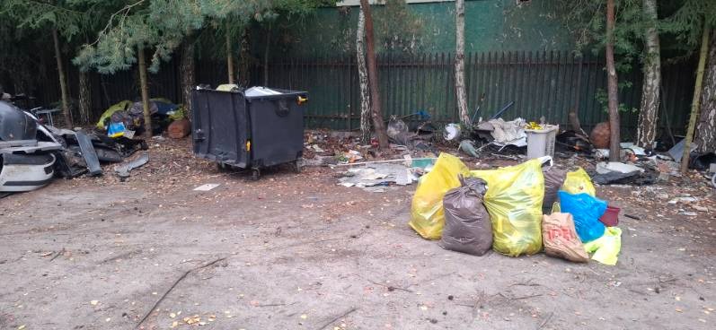 Kontener na śmieci i worki z odpadami na zewnątrz, wśród drzew i ogrodzenia. Widoczne elementy samochodowe i inne odpady.
