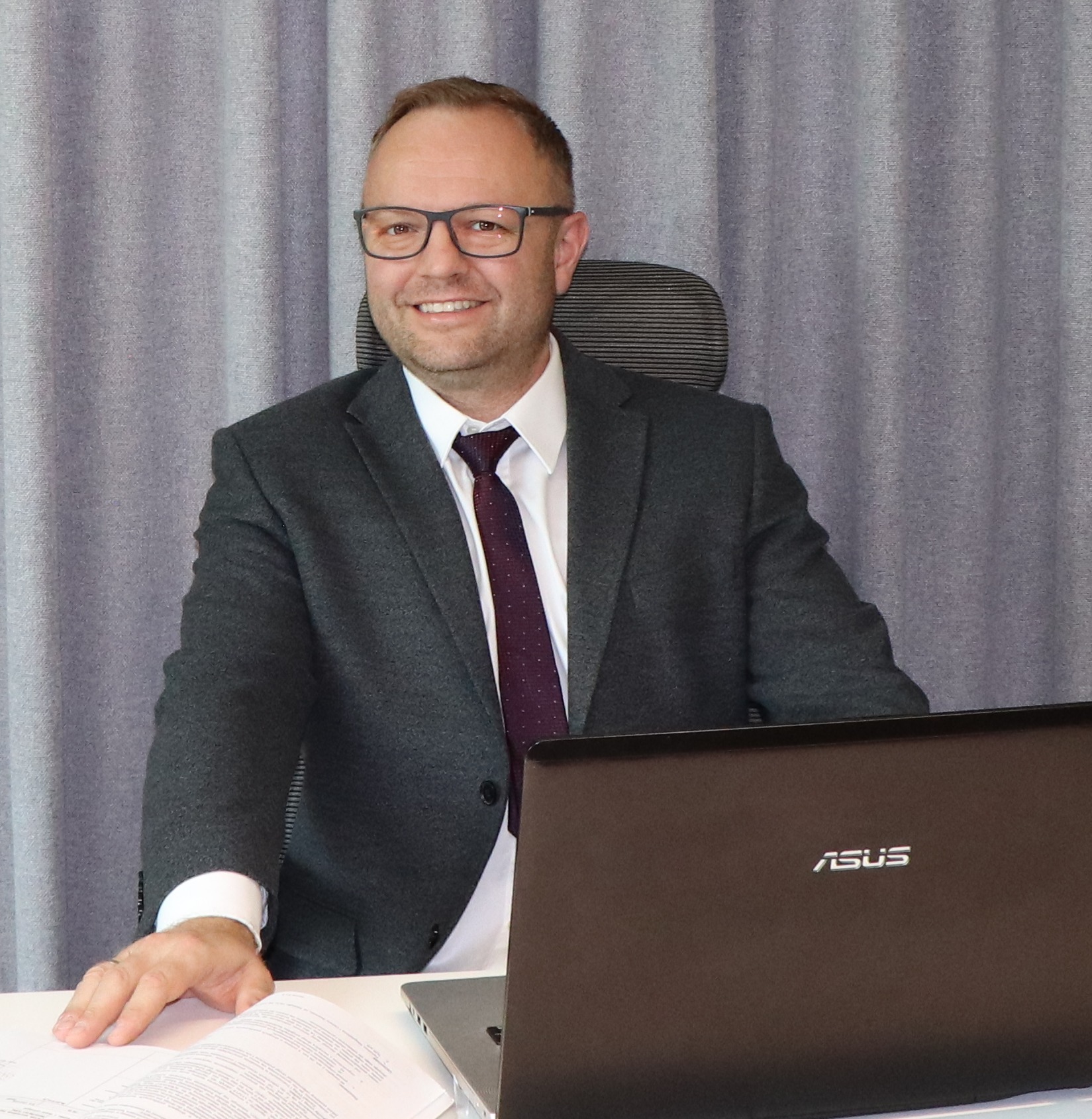 Uśmiechnięty administrator nieruchomości w garniturze siedzi za biurkiem z laptopem Asus, przeglądając dokumenty na tle szarej zasłony.