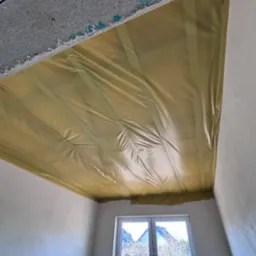 Żółta folia zabezpieczająca na suficie w trakcie remontu, widok z dołu, okno w tle. Zabezpieczenie przed malowaniem lub szpachlowaniem.