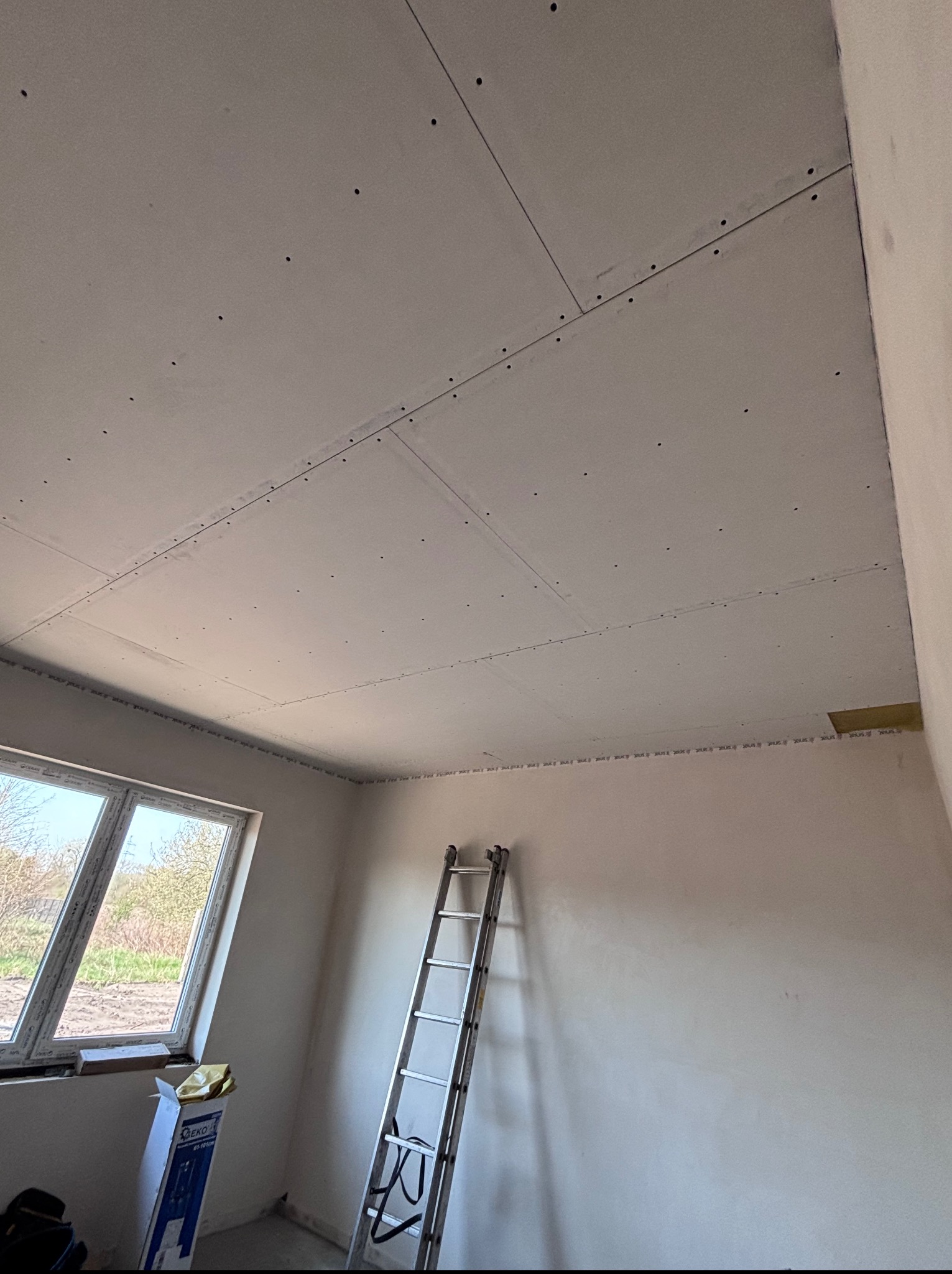Montaż płyt karton gips na suficie w jasnym pomieszczeniu. Widoczna drabina oparta o ścianę, okno z widokiem na zewnątrz i kartonowe opakowanie na podłodze.